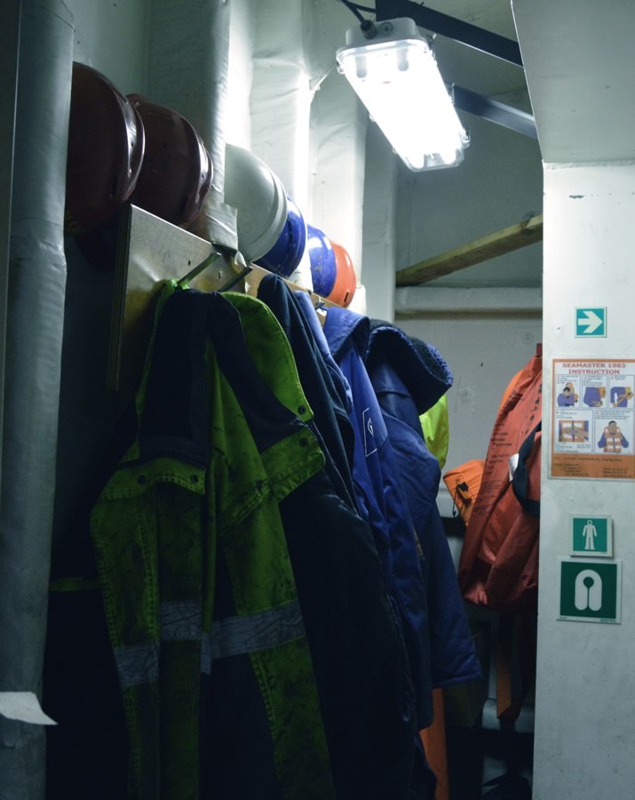 Work jackets and helmets hanging in a ship's gear room under a fluorescent light